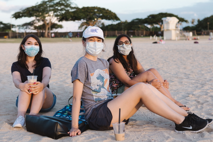 Three young people wearing masks and sitting in the sand together