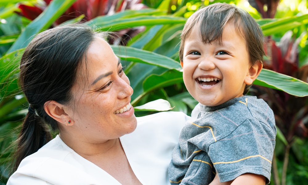 Smiling woman holding a cheerful young child outdoors, surrounded by vibrant green plants.