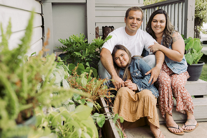 A family sits together on their porch, smiling, surrounded by plants.